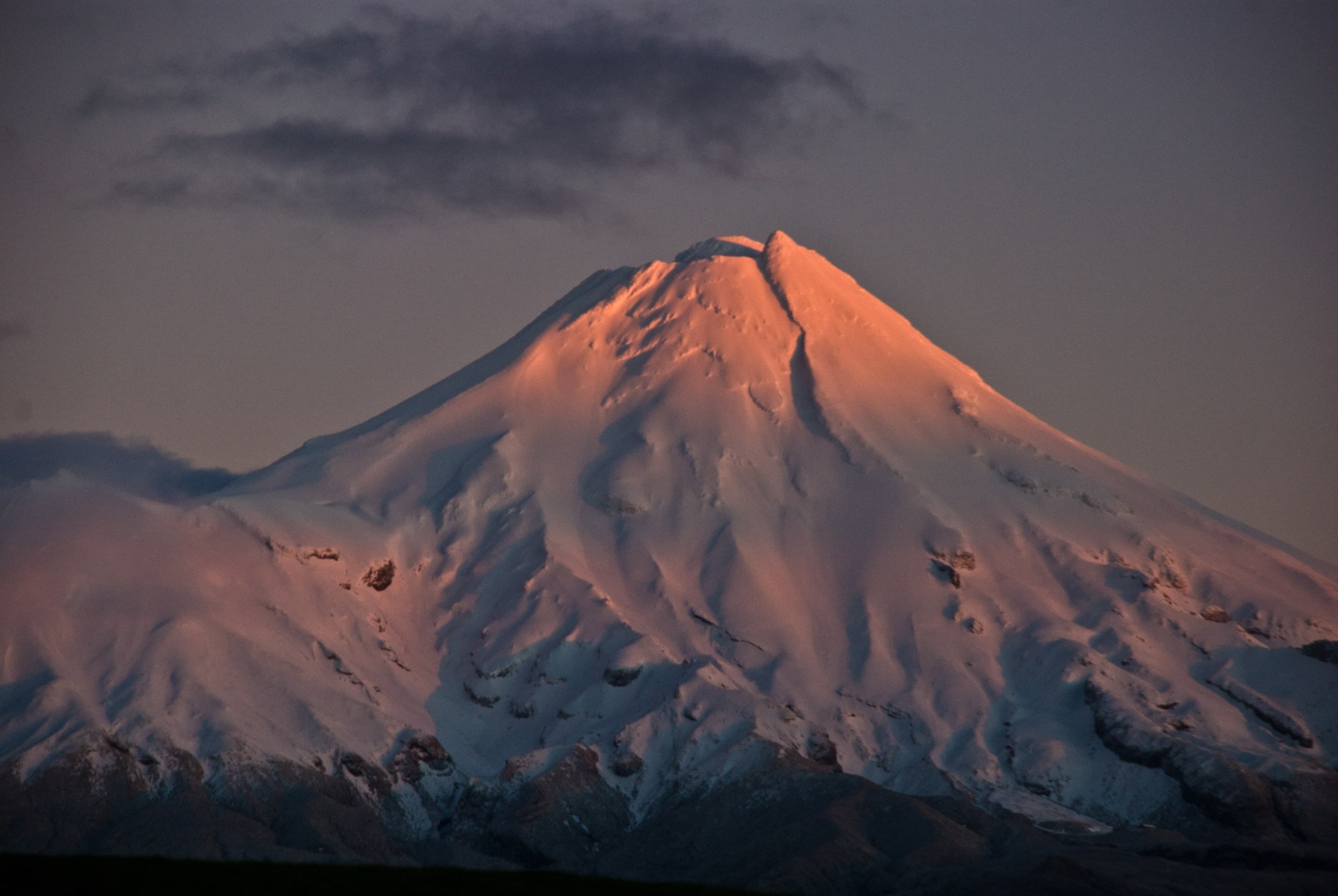 Snow-covered summit of Mount Taranaki lit orange at daybreak beneath dark clouds